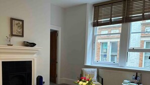 Ein Wohnzimmer mit Kamin, Couch, Tisch mit Blumen und einem Fenster mit Jalousien.