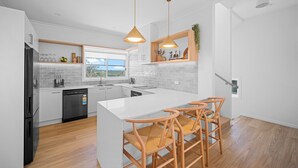A modern kitchen with white cabinetry, a central island with bar stools, and pendant lighting. - The Lookout – Resort Living with Panoramic Views Over Diamond Beach (Diamond Beach)