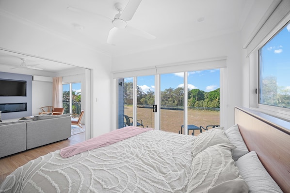 A modern bedroom with a large bed, a TV, a sofa, and a balcony with a view of greenery.