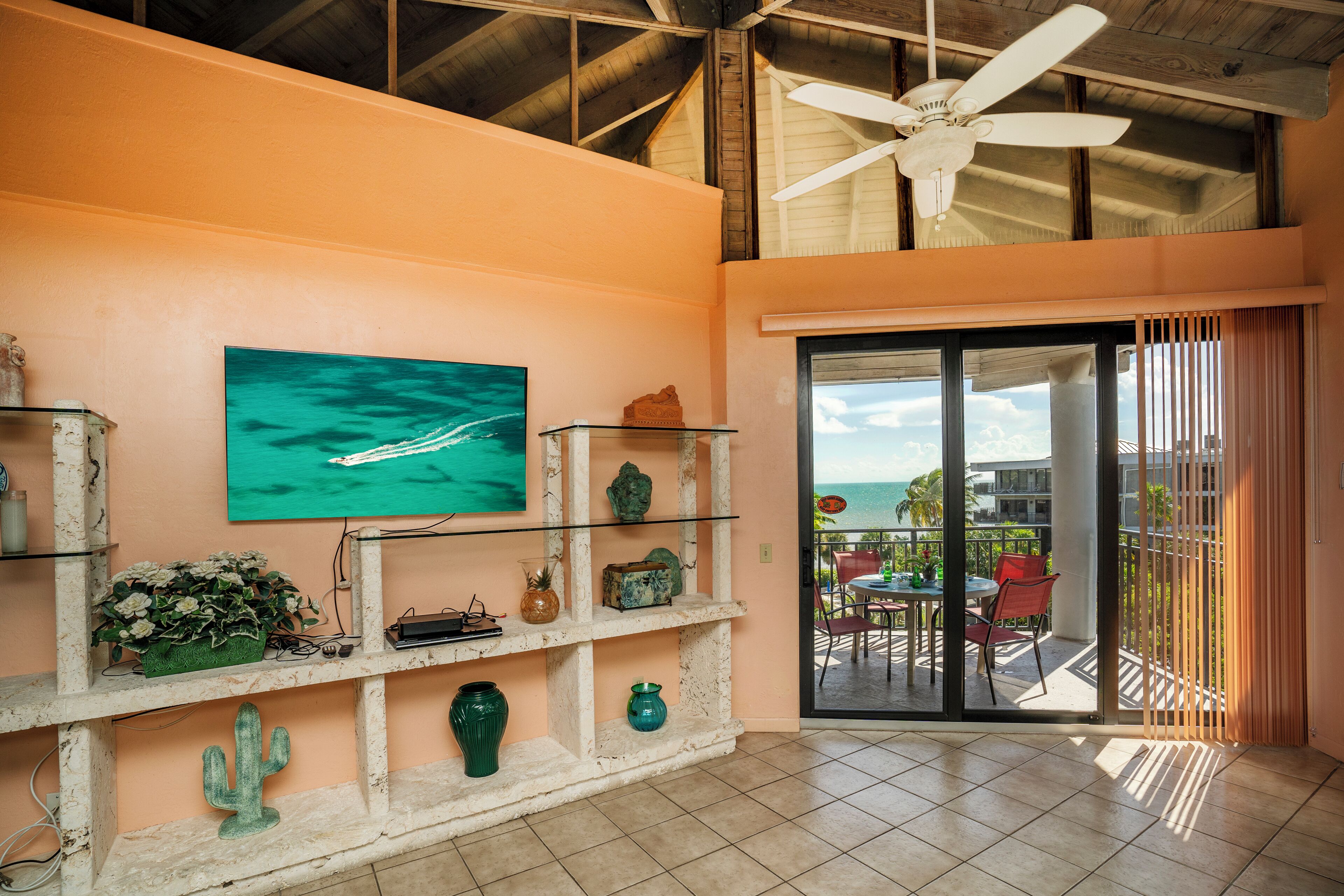 A room with a ceiling fan, a large window with a view of the ocean, a cactus decoration, and a shelf with various items.