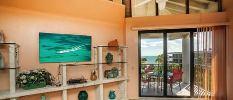 A room with a ceiling fan, a large window with a view of the ocean, a cactus decoration, and a shelf with various items.