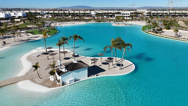 An aerial view of a resort with a large, curved swimming pool, palm trees, and a distant view of a cityscape. - Designer Villa with Heated Pool on Santa Rosalia Lake Resort (Torre-Pacheco)