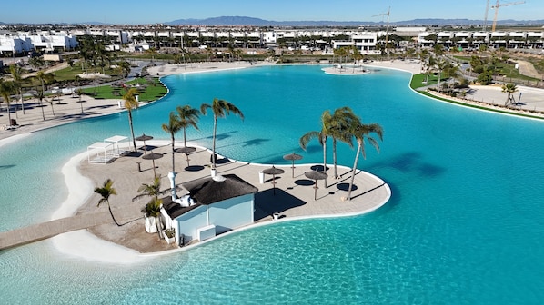 An aerial view of a resort with a large, curved swimming pool, palm trees, and a distant view of a cityscape. - Designer Villa with Heated Pool on Santa Rosalia Lake Resort (Torre-Pacheco)