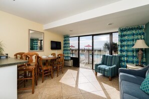 Ein Zimmer mit Esstisch, Stühlen, einer Couch, einem Fernseher und einem Balkon mit Sonnenschirmen.