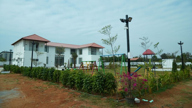 A building with a red roof, surrounded by a fenced garden with various plants and a swing set.