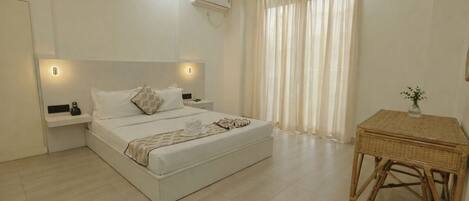 A modern hotel room with a large bed, bedside tables, an air conditioning unit, a window with curtains, and a wicker side table.