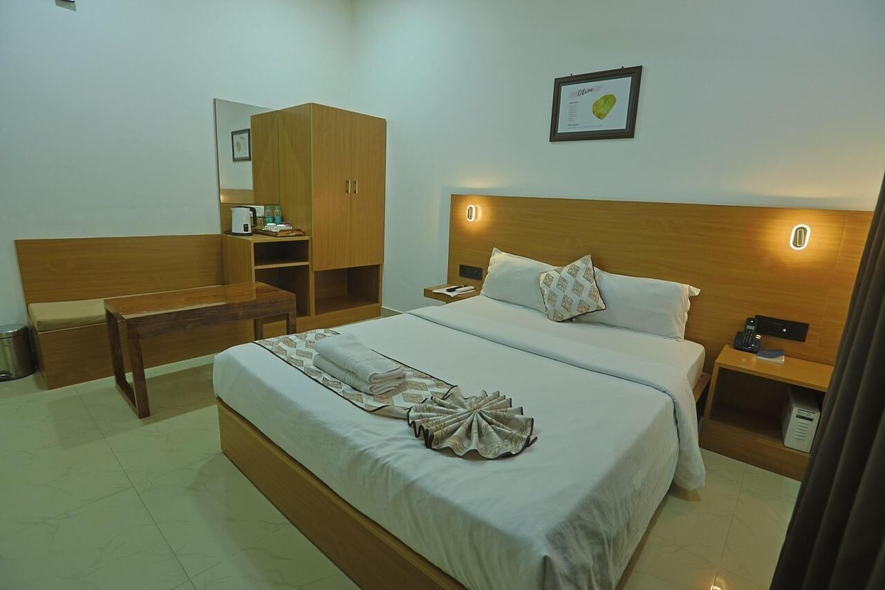 A hotel room with a large bed, a wooden wardrobe, a bedside table with a lamp, and a small desk with a chair.