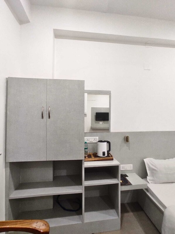 A bedroom with a grey wardrobe, a bedside table with a kettle, a power outlet, and a bed with white bedding.