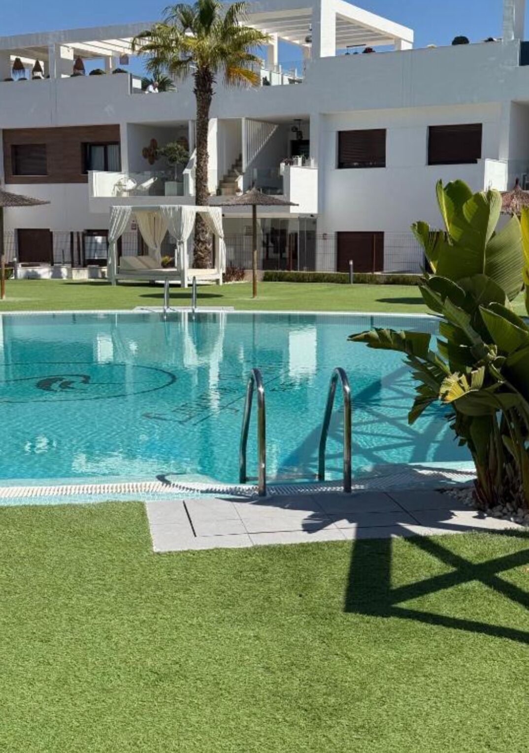 Ein Swimmingpool mit klarem, blauem Wasser, umgeben von künstlichem Rasen, mit einem weißen Gebäude mit Balkonen im Hintergrund.