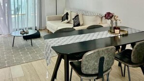 Een moderne woonkamer met een bank, eettafel, stoelen, een hanglamp en een groot raam met dunne gordijnen.