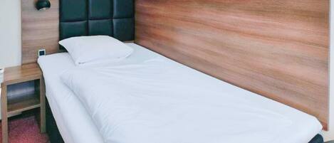 A single bed with a wooden headboard, white bedding, a bedside table, and a red carpet.