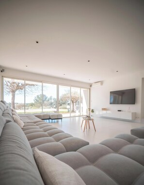 Ein modernes Wohnzimmer mit einem großen Ecksofa, einem flachbildfernseher an der Wand und einem großen Fenster mit Blick ins Freie.