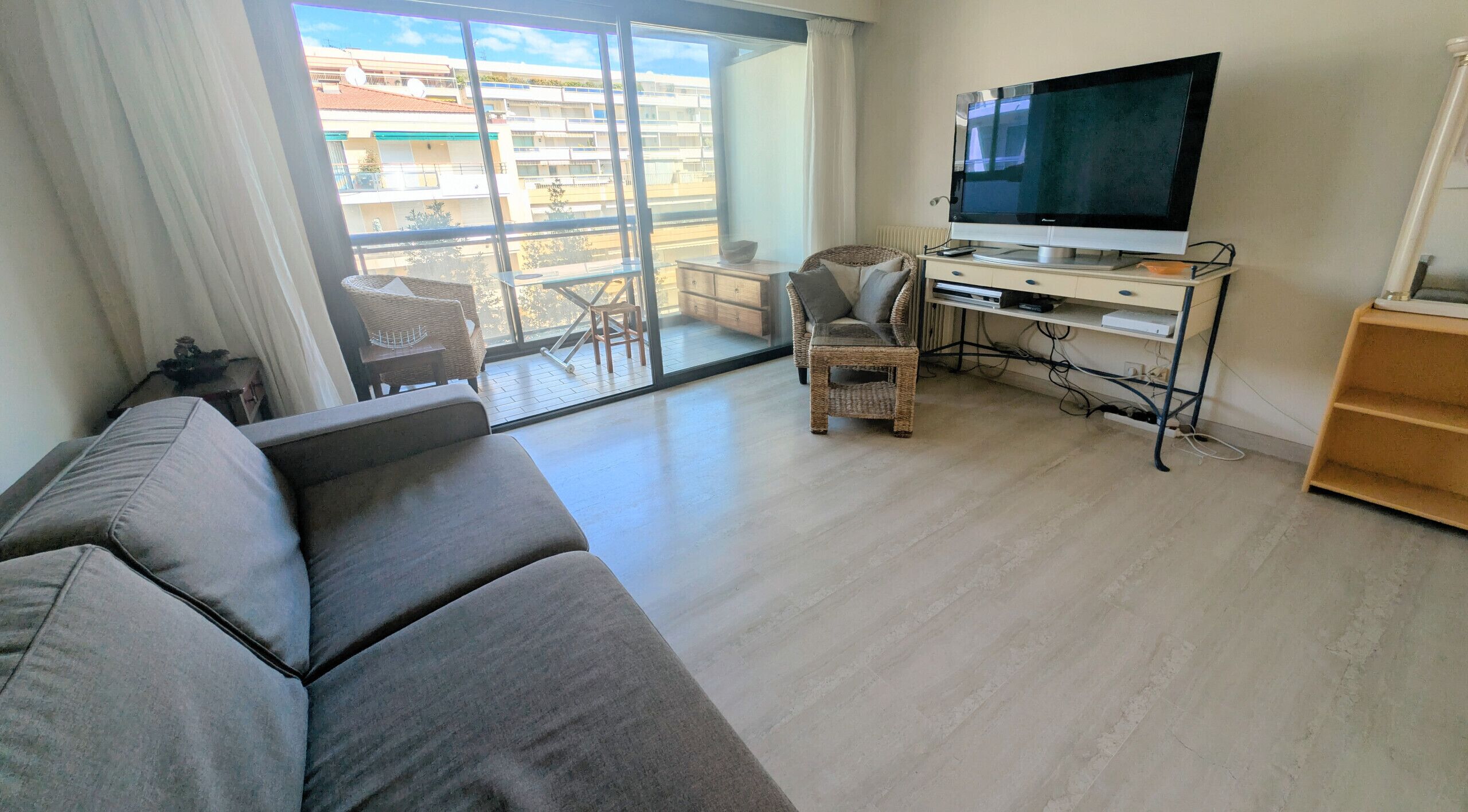 Ein modernes Wohnzimmer mit einer Couch, einem Fernseher auf einem Schrank, einem Korbsessel und einem Balkon mit Blick auf Gebäude.
