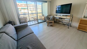 Ett modernt vardagsrum med en soffa, en tv på ett tv-bord, en fåtölj i rotting och en balkong med utsikt över byggnader.
