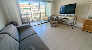A modern living room with a sofa, a television on a stand, a wicker chair, and a balcony with a view of buildings. - Ref JDC by Palmes d Or Properties (Cannes)
