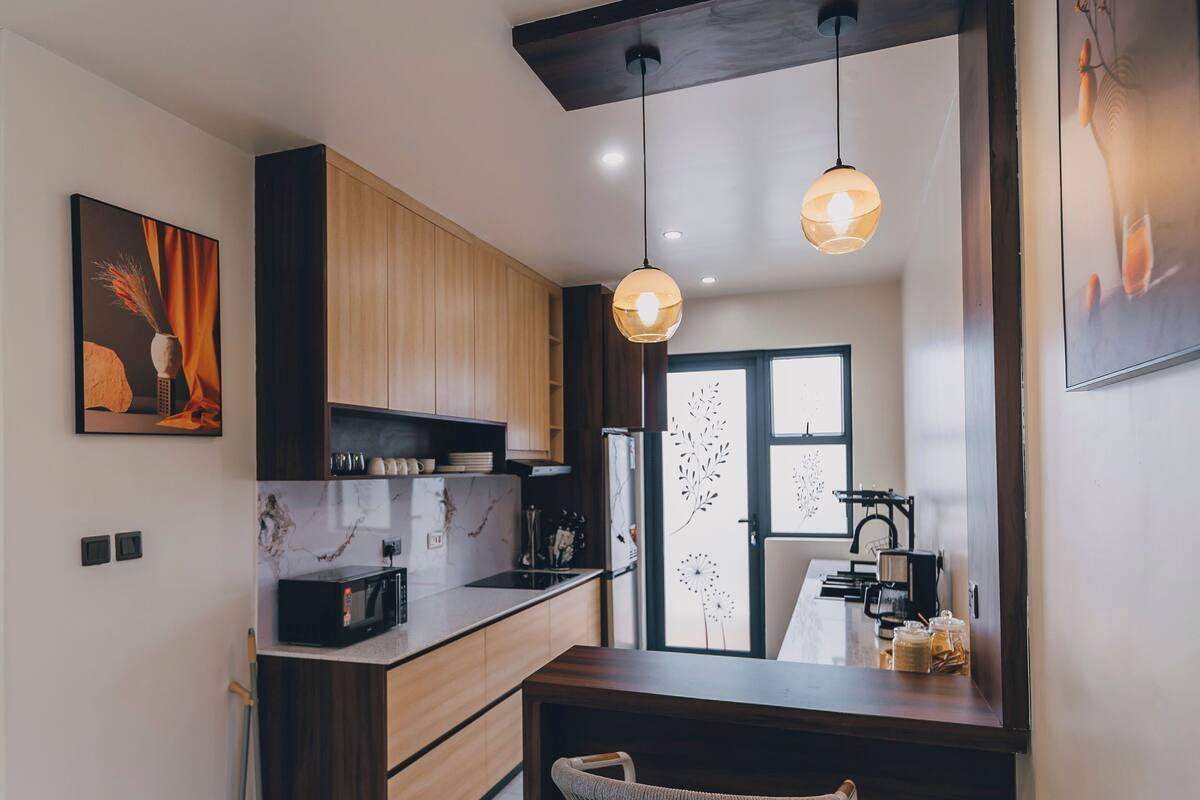 A modern kitchen with wooden cabinets, a marble countertop, a hanging light fixture, and a dining area with a chair.