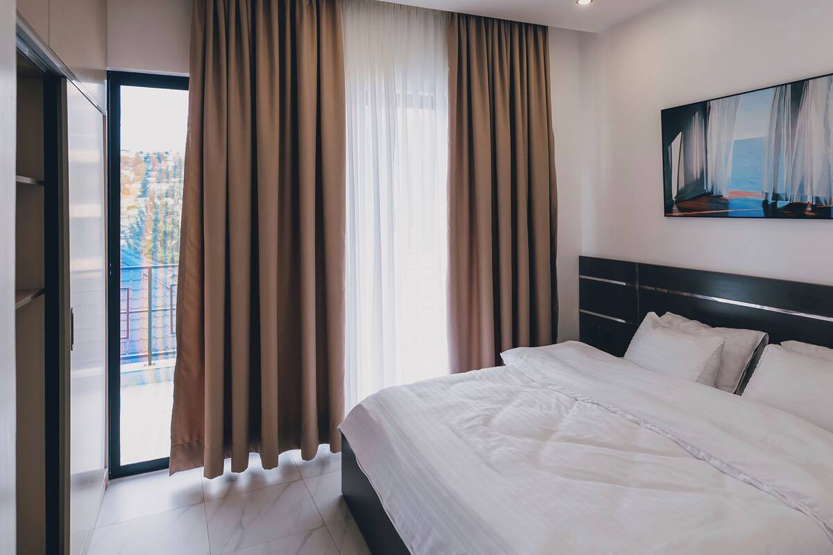 A modern hotel room with a large bed, a sliding glass door to a balcony, and a painting on the wall.