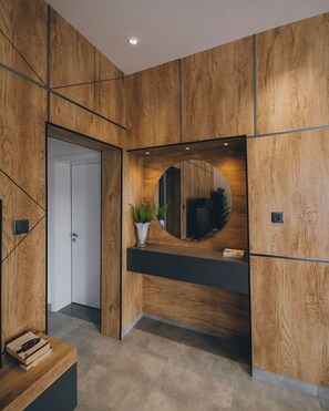 Un interno moderno caratterizzato da pannelli di legno, uno specchio centrale, un piano d'appoggio scuro e una porta che dà su un'altra stanza.