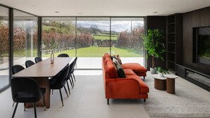 A modern living room with a large glass wall, a sofa, a dining table with chairs, and a television. - Woldside - Luxury Property Sleeps 10 (York)