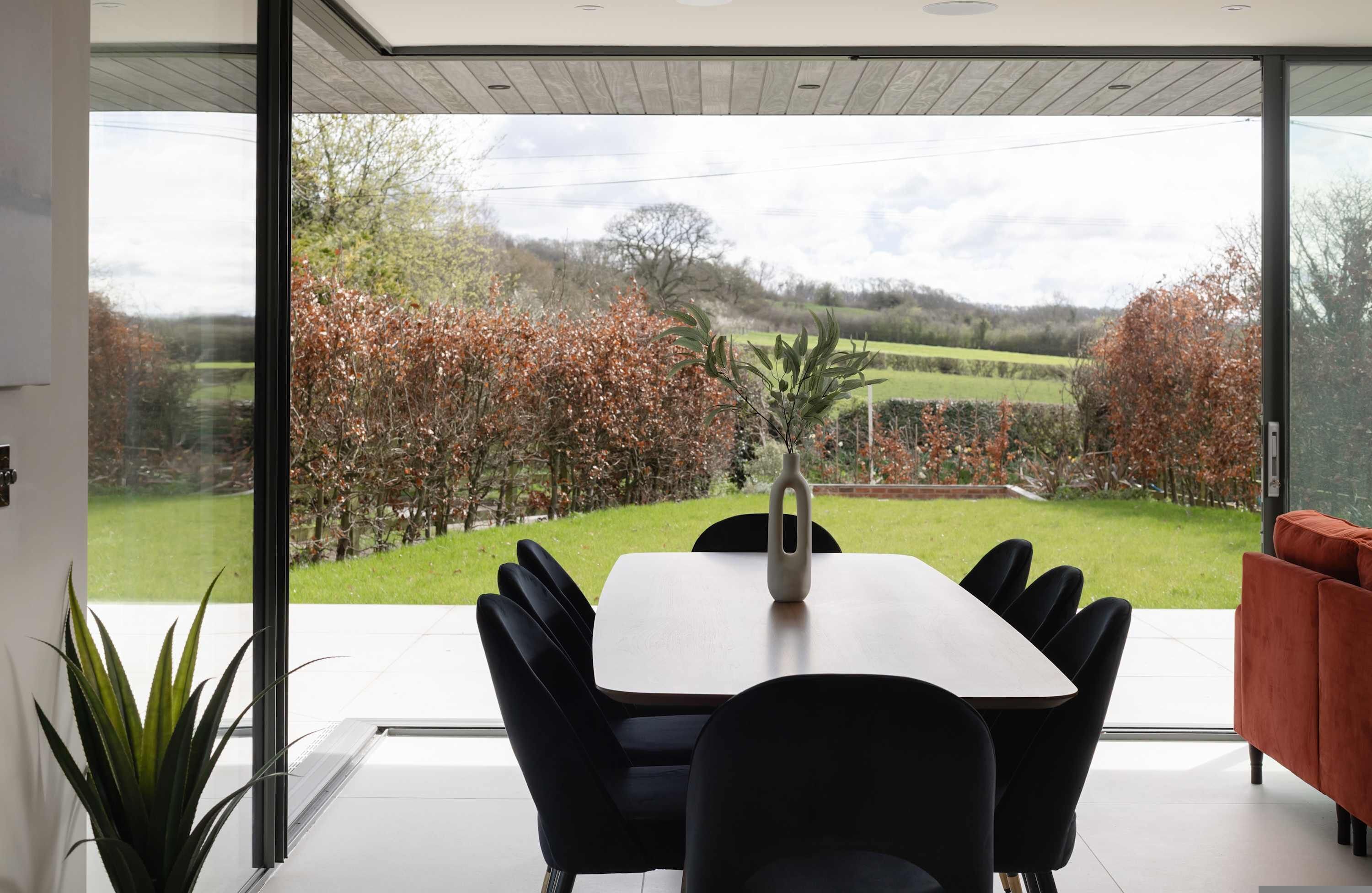 A modern dining area with a table and chairs, large windows with a view of a garden and countryside.