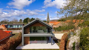 A modern house with a glass facade, a brick lower level, and a visible church spire in the background. - Woldside - Luxury Property Sleeps 10 (York)