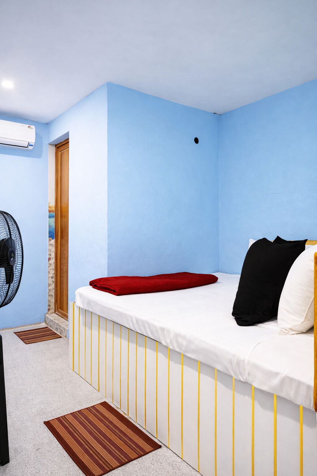 A room with a bed, a standing fan, a door, and a wall-mounted air conditioner.