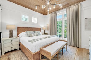 A bedroom with a large bed, bedside tables, a bench, a ceiling light fixture, and a window with curtains. - Comin' In Hot: 5 bed new build across from Carillon Chapel & Village Green (Panama City Beach)