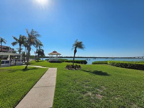 Ein gepflegter Rasen mit einem Gazebo, Palmen und freiem Blick auf das Wasser mit Booten.