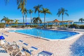 A swimming pool with lounge chairs and umbrellas, surrounded by palm trees and a view of the water. - Waterfront Oasis on Isla Del Sol in Bahia Del Mar (St. Petersburg)