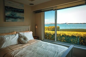 A bedroom with a large window, a bed with pillows, a nightstand, and a painting on the wall. - Waterfront Oasis on Isla Del Sol in Bahia Del Mar (St. Petersburg)