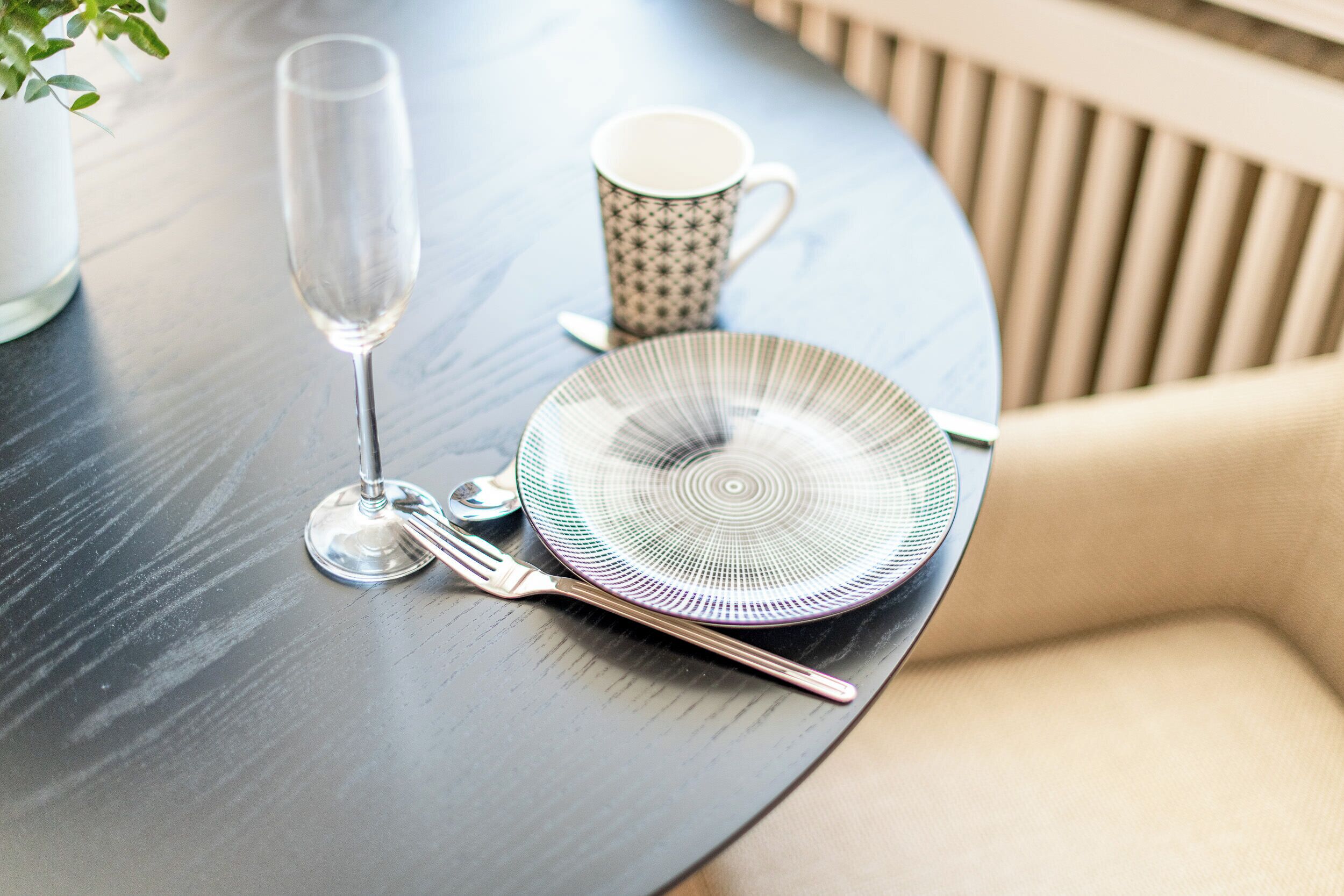 A round dining table with a clear glass, a patterned mug, a plate with a radial design, and cutlery.