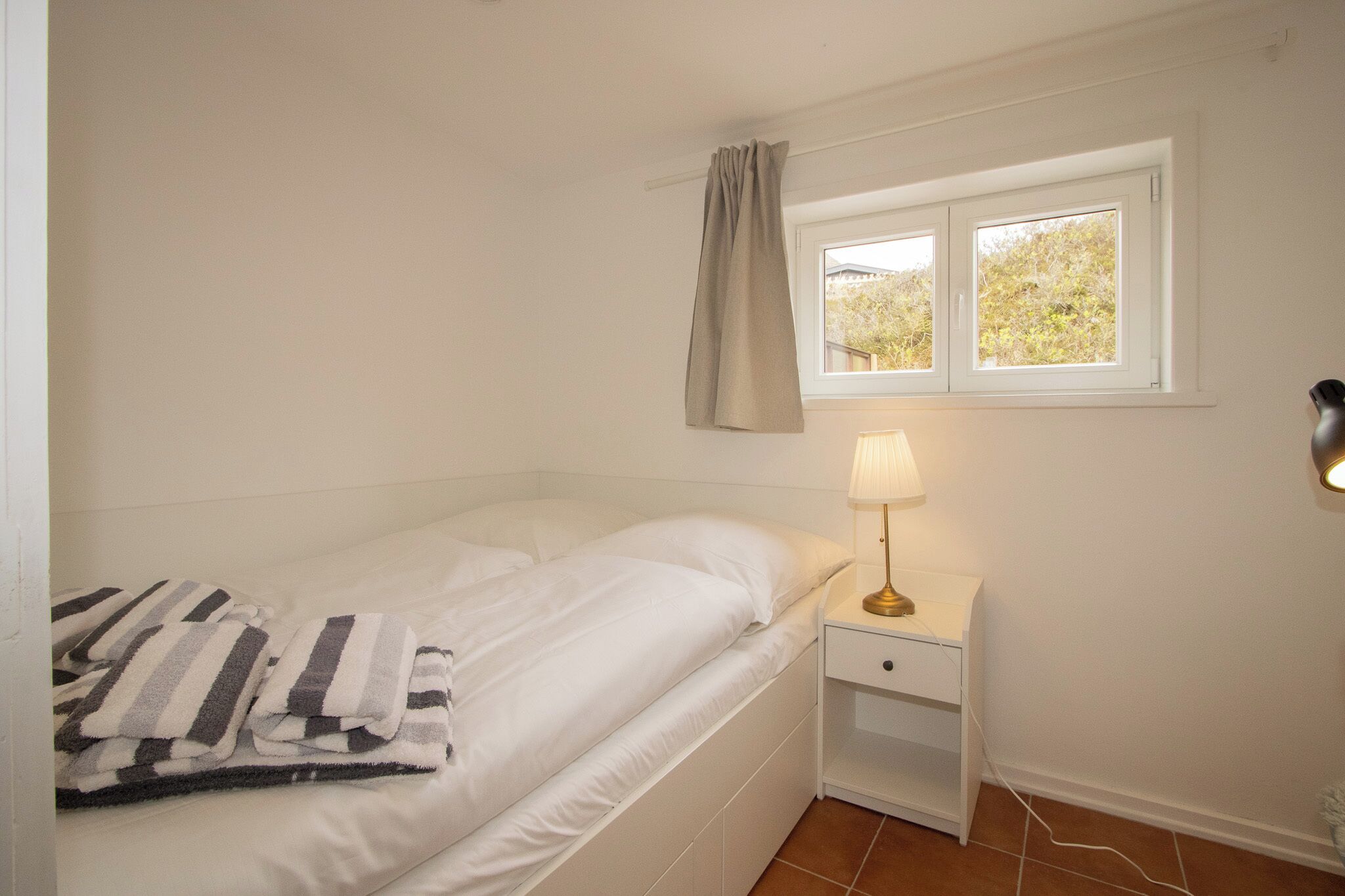 A single bed with a white mattress and pillows, a bedside table with a lamp, a window with a view of trees, and a curtain.