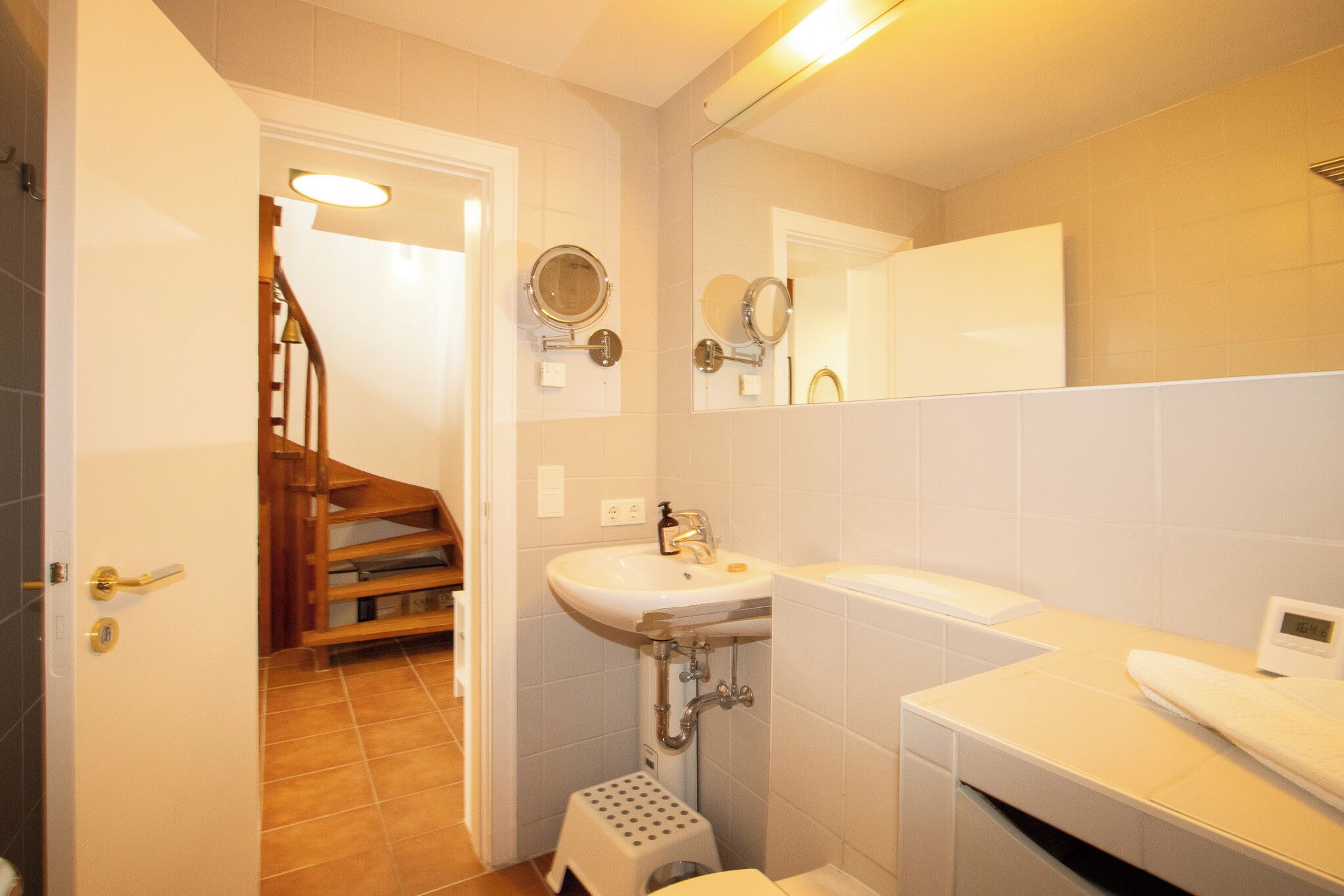 Un baño con lavabo, espejo y retrete. Se ve una escalera de madera que conduce a otra habitación.