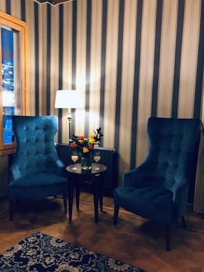 Ein Zimmer mit Streifenmuster-Tapete, zwei blaue Sessel, ein kleiner Tisch mit Blumenvase und eine Lampe.