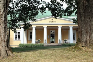 Um edifício com pórtico sustentado por colunas, uma placa com a inscrição 'BACKBY' e um gramado bem cuidado na frente.