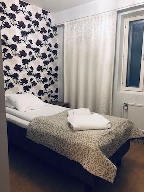 Ein Einzelbett mit einem gemusterten Überwurf, ein Nachttisch, ein Fenster mit Blick und eine Wand mit floralem Tapetenmuster.