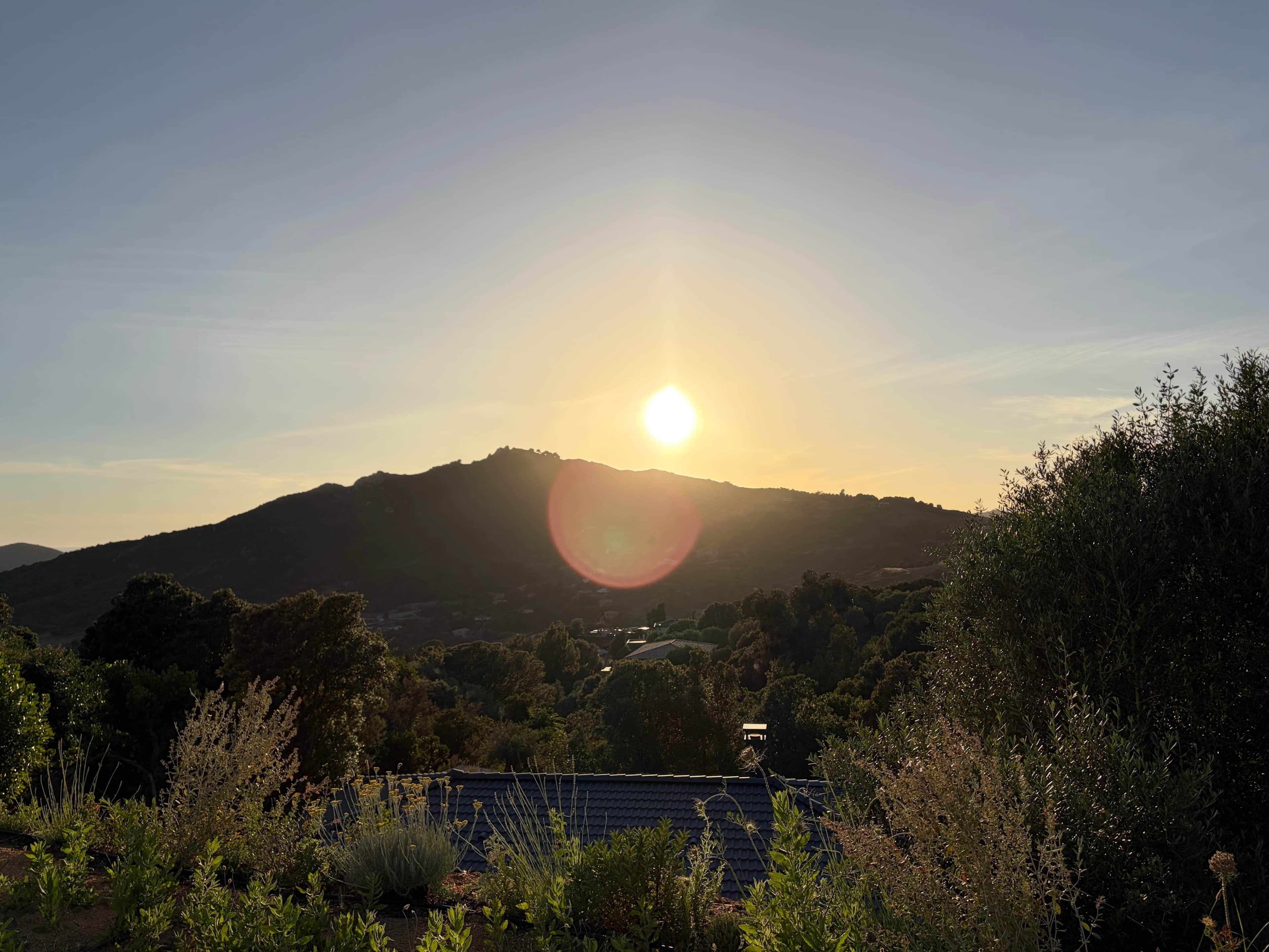 Un paysage où le soleil se couche derrière une colline, entouré de verdure et où se trouve un bâtiment avec un toit sombre.