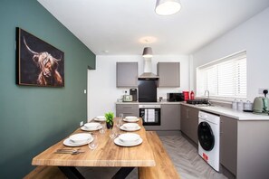 A modern kitchen with a dining table set for four, a washing machine, and a large window with blinds.