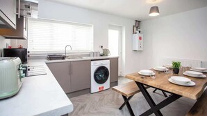 A modern kitchen with a built-in washing machine, a dining area with a wooden table set for four, and a large window providing natural light. - Vacant nests | The Cowpen | 3BR | Parking & Wi-Fi (Blyth)