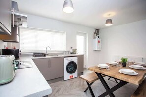 A modern kitchen with a built-in washing machine, a dining area with a wooden table set for four, and a large window providing natural light.