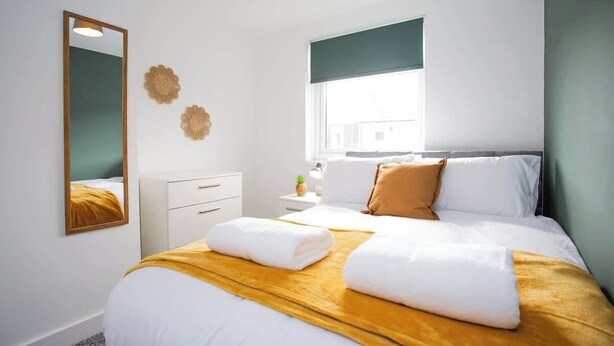A hotel room with a bed, a nightstand, a mirror, and a window with a view of buildings. - Vacant nests | The Cowpen | 3BR | Parking & Wi-Fi (Blyth)