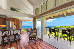 A kitchen with a view of the ocean, a dining area with chairs, and a balcony with a table and chairs. - Pali Ke Kua #231 Serene Ocean Views, Walk to Beach (Princeville)
