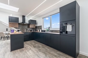 A modern kitchen with dark cabinetry, a built-in oven, and a window overlooking the sky. - Luxury 2 bed Penthouse with Views over Central London (Free Parking) (Uxbridge)