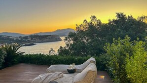 A wooden deck with a wicker sofa and two black stools, overlooking a body of water and hills. - Crete Beachfront Coconut Gem With Seaviews Pool (Apokoronas)