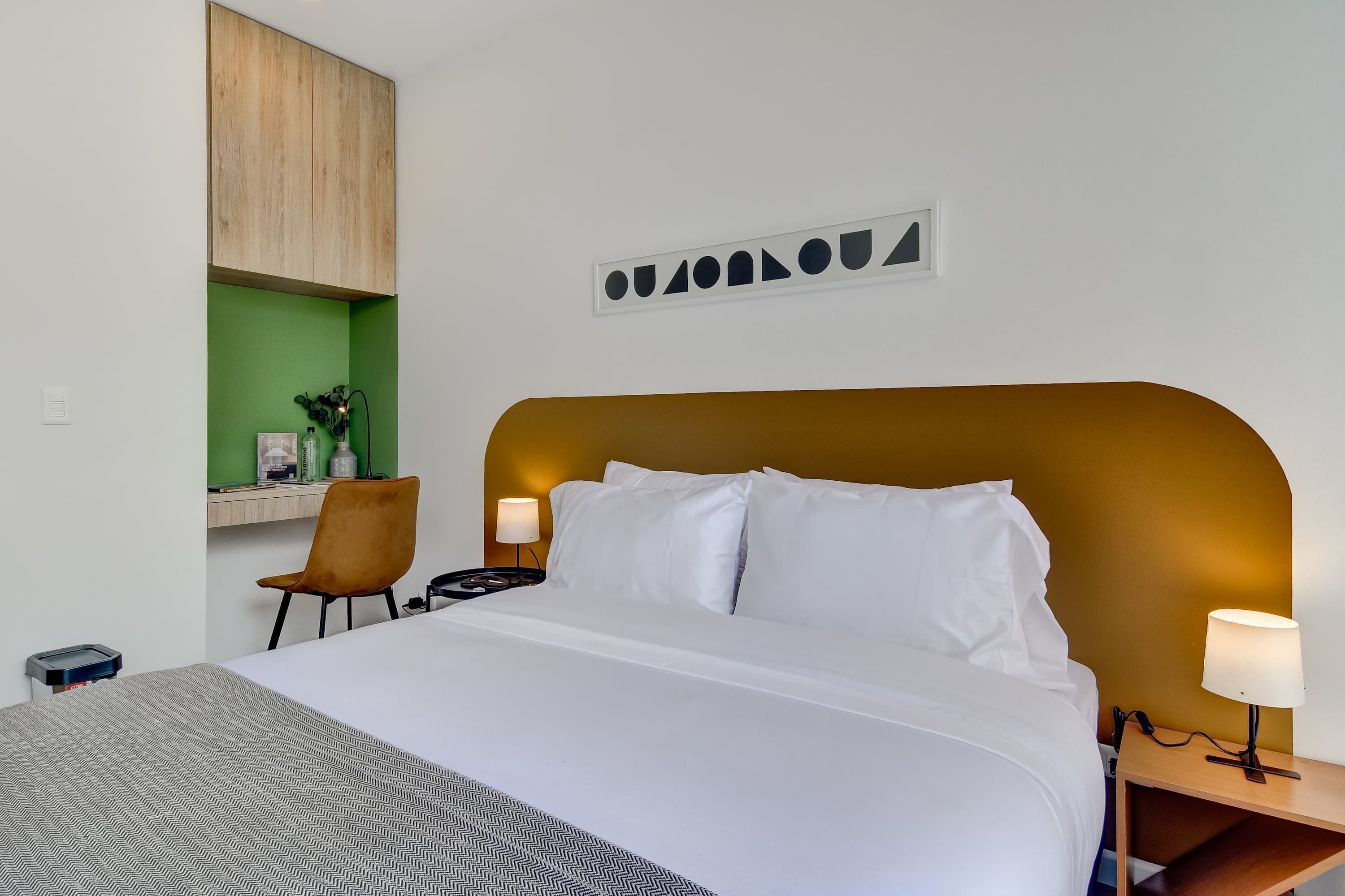 A hotel room with a bed, a desk, a chair, a nightstand, and a wall-mounted cabinet.