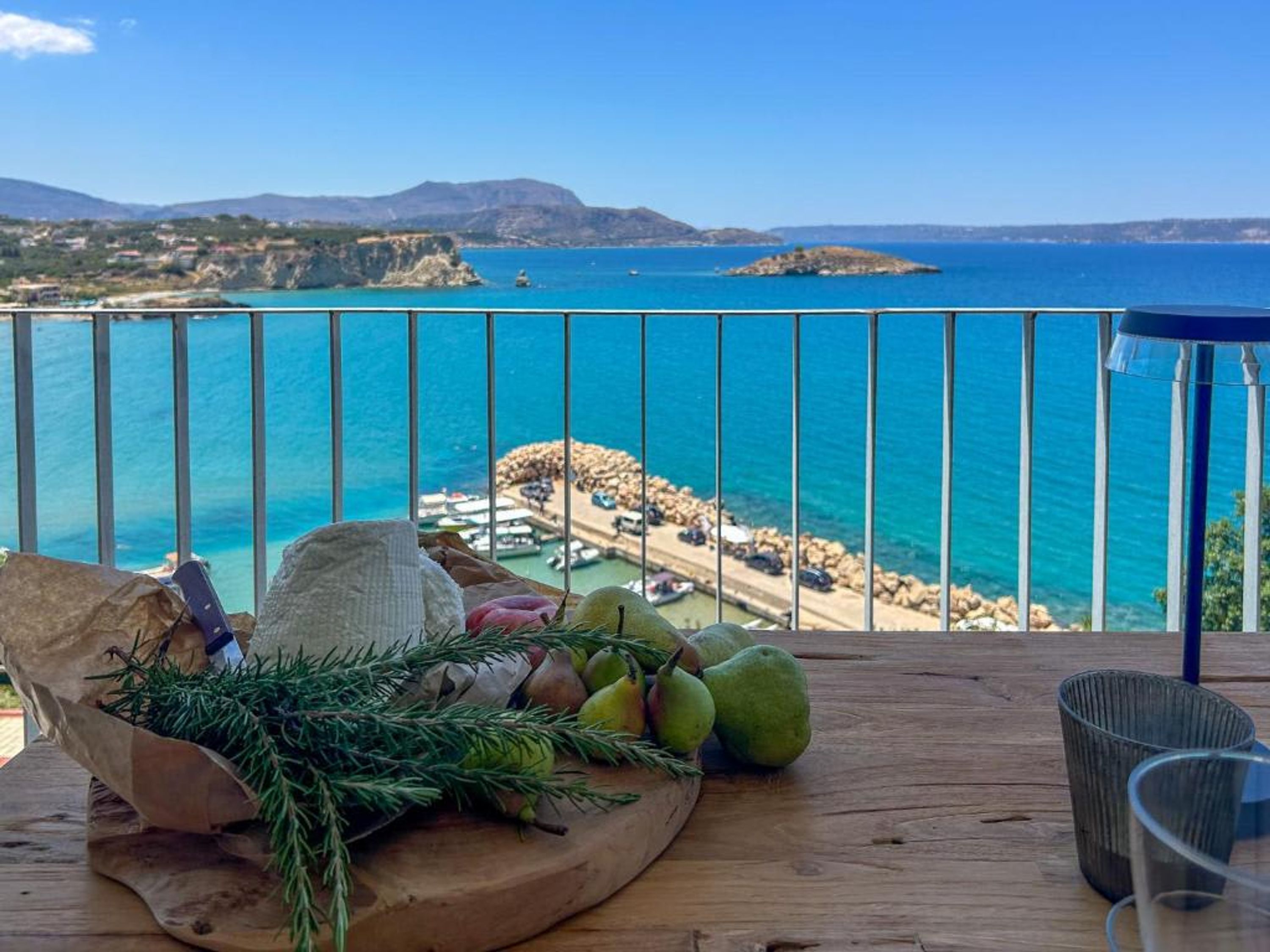 Ein Holztisch mit Früchten, Nüssen und einem Zweig, mit Blick auf die Küste, wo Boote und felsiges Gelände zu sehen sind.