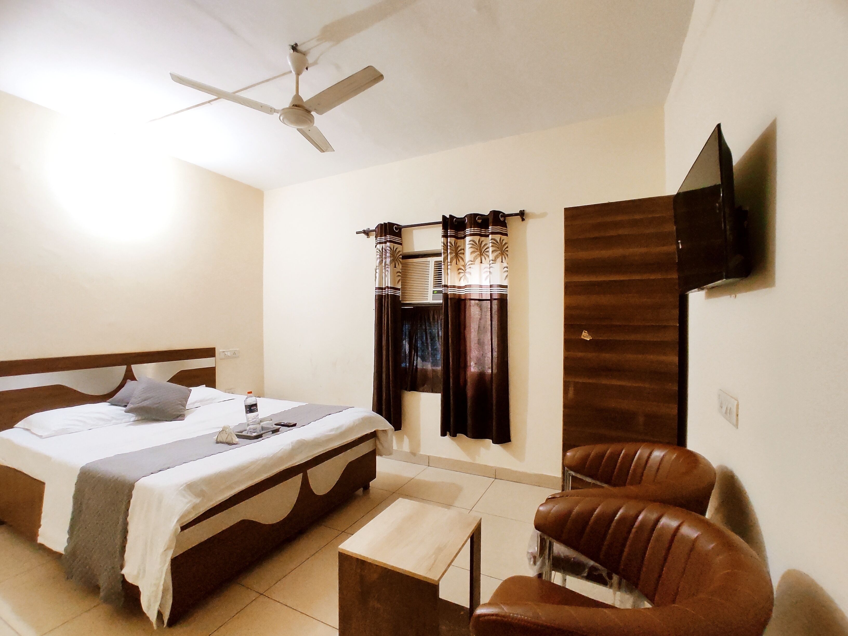 A bedroom with a bed, a chair, a small table, a ceiling fan, curtains, and a TV mounted on the wall.