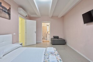 Quarto com cama, sofá, televisão e banheiro visível por uma porta aberta.