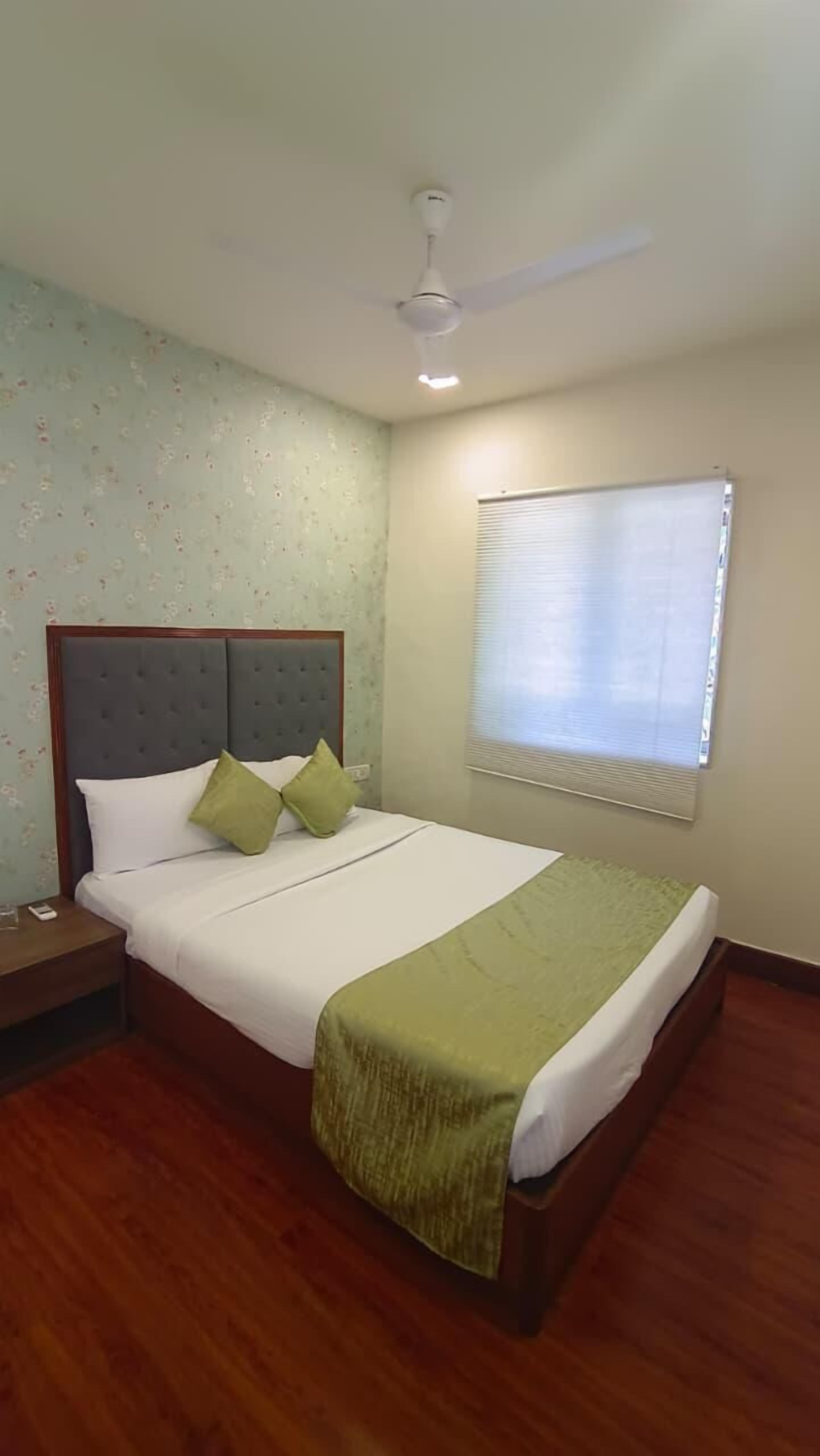 A hotel room with a bed, a nightstand, a ceiling fan, a window with blinds, and a floral wallpaper pattern.