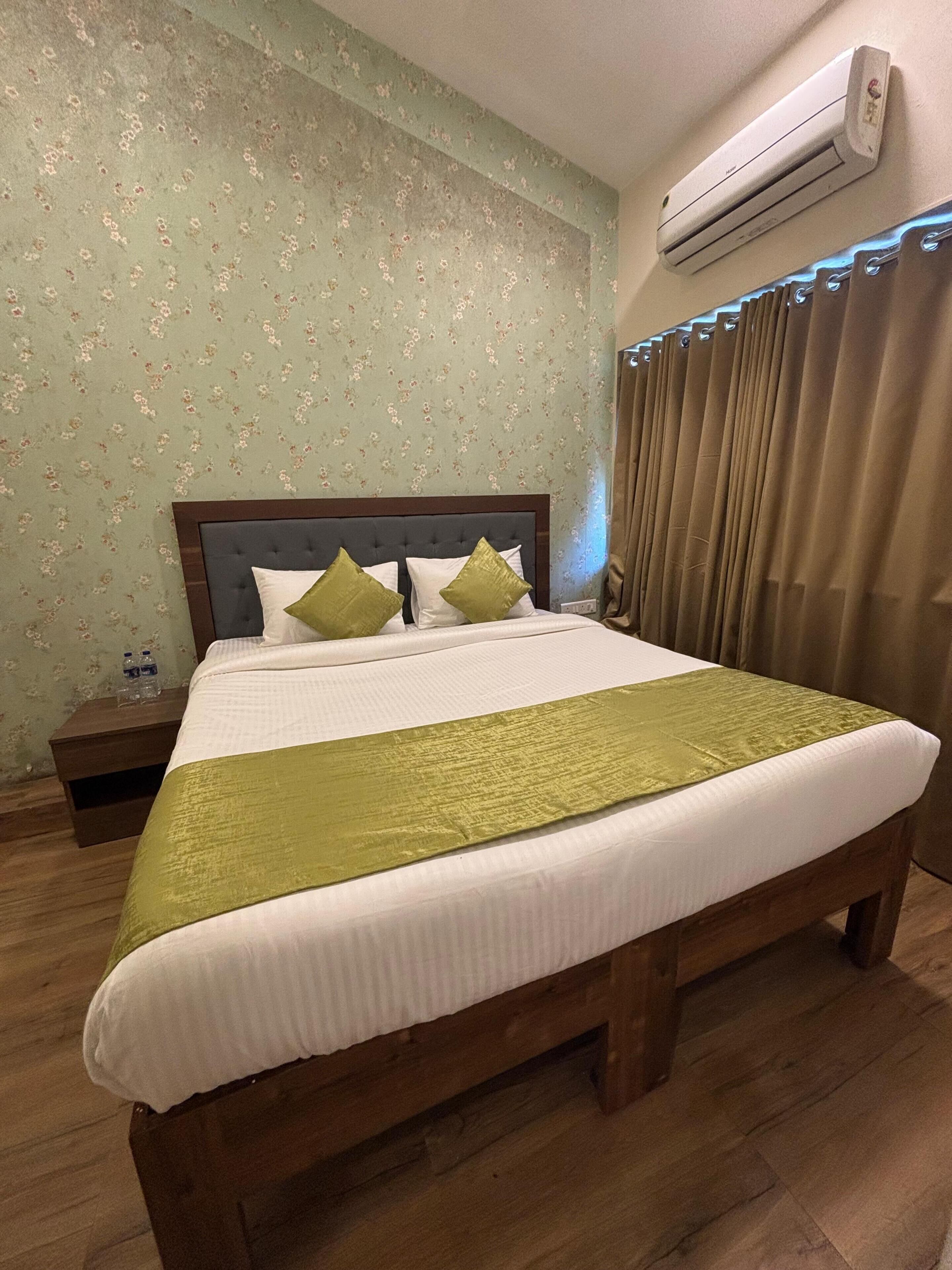 A neatly made bed with a green bedspread, a wooden headboard, and two pillows. There is a nightstand with a bottle and glasses, and an air conditioning unit on the wall.